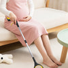 A woman wearing a pink dress is using a yellow and black foldable reacher grabber tool on a light-colored carpet.