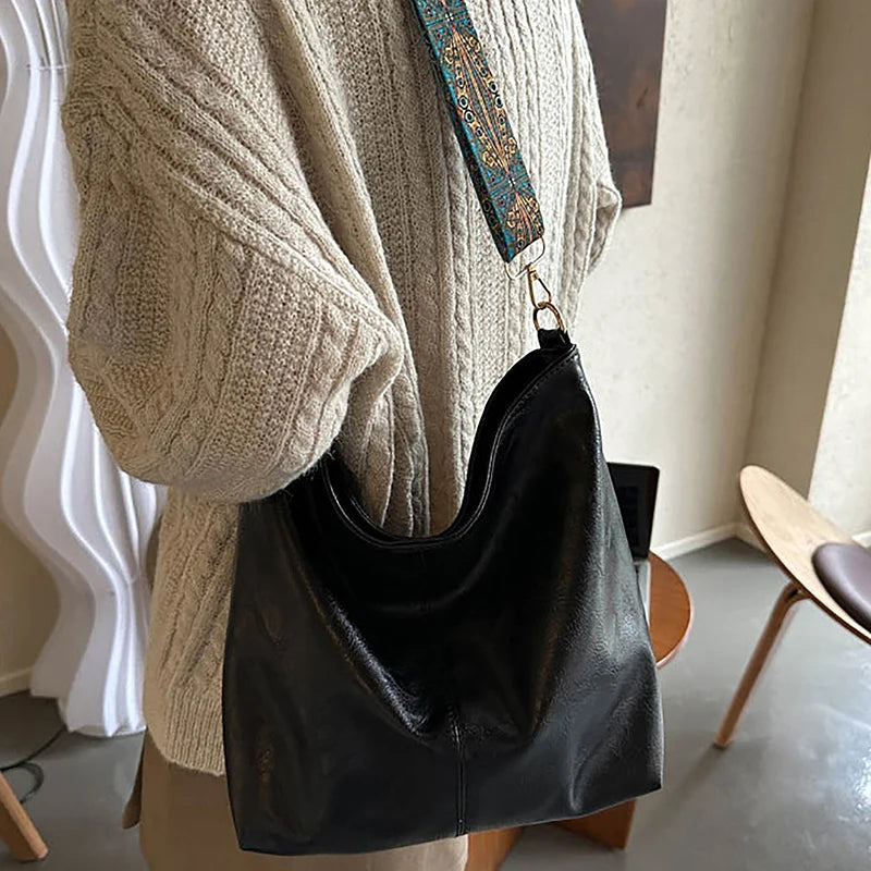 Sac à main en cuir noir avec une bretelle géométrique large, suspendu d'un pull tricoté beige.