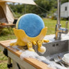 A yellow 3D sponge holder with a blue sphere is positioned over a kitchen sink.
