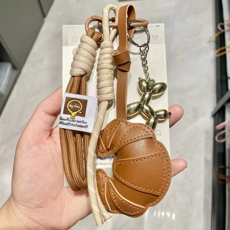 A braided leather croissant keychain with a bag pendant is displayed against a white background.