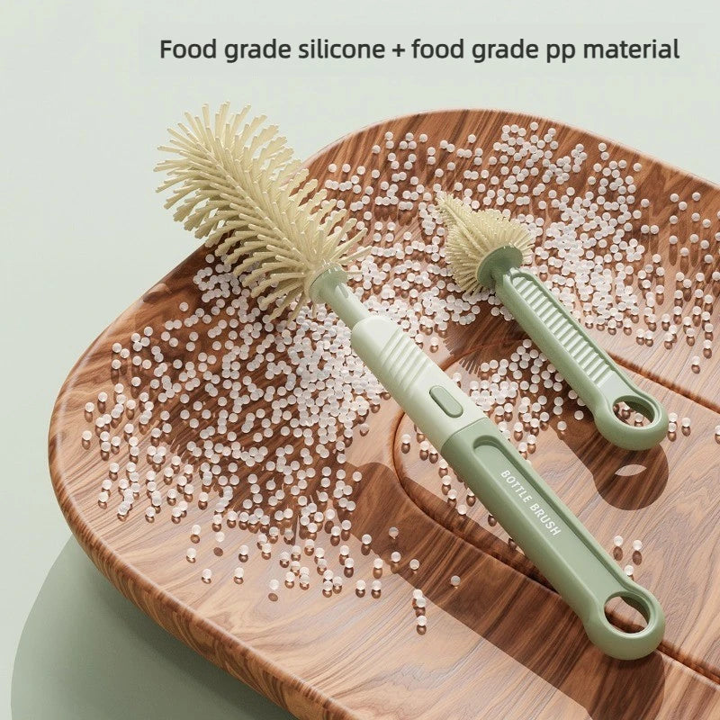A silicone baby bottle brush set with a light green handle and bristles is shown resting on a wooden surface surrounded by sm