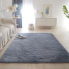A fluffy blue rug is placed on a light wood floor in a bright, minimalist room with white furniture and natural light.