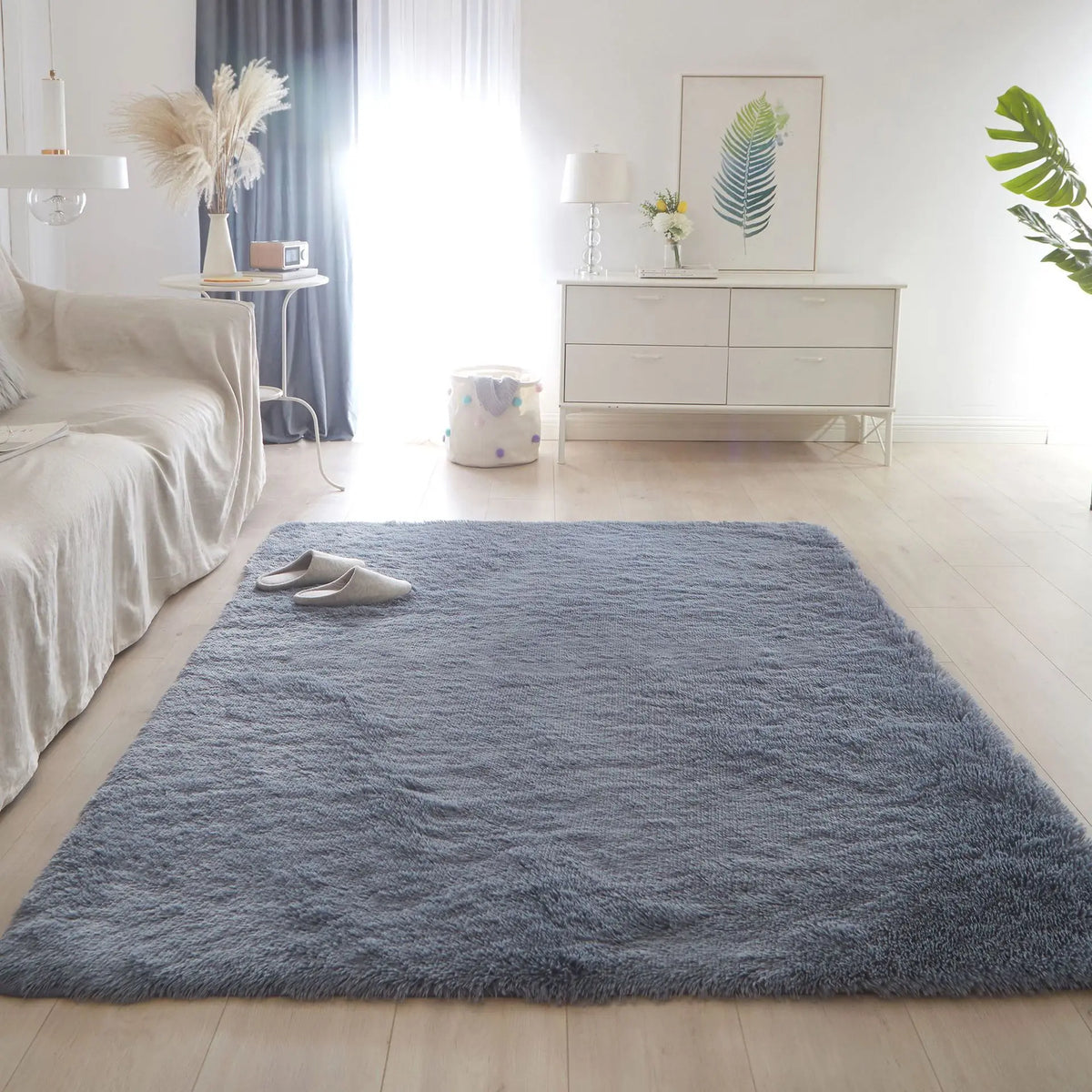 A fluffy blue rug is placed on a light wood floor in a bright, minimalist room with white furniture and natural light.