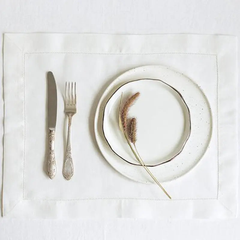 A set of six linen hemstitch napkins displayed on a white cloth with silverware and a plate.