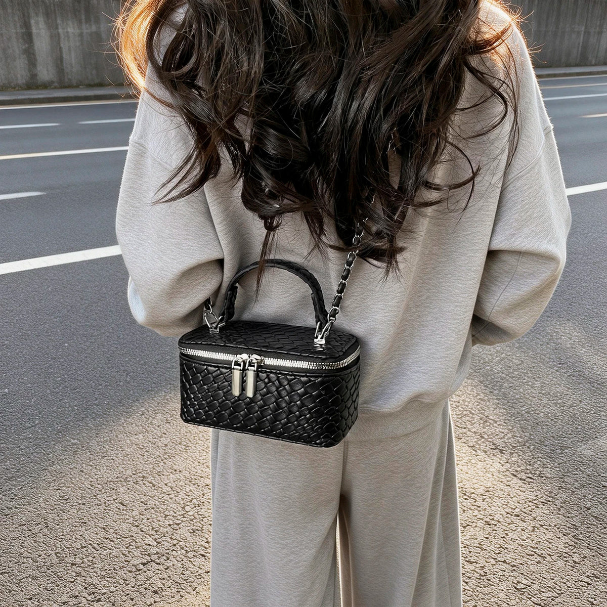 A woman is holding a black woven PU leather crossbody bag with a silver chain against a gray background.