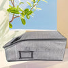A gray foldable fabric storage box with a zipper is shown on a light surface next to a green branch.