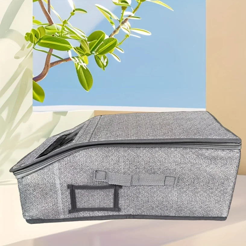 A gray foldable fabric storage box with a zipper is shown on a light surface next to a green branch.