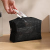 A hand is opening a black lace mesh makeup bag with a clear glass bottle inside.