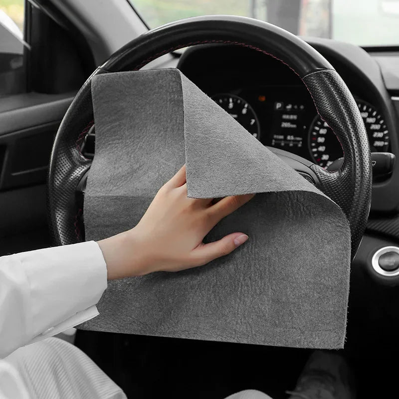 A hand is holding a gray microfiber cleaning cloth against the steering wheel of a car.