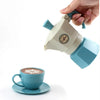 A hand is holding a light blue and cream colored coffee maker or teapot, with a matching cup and saucer visible below.