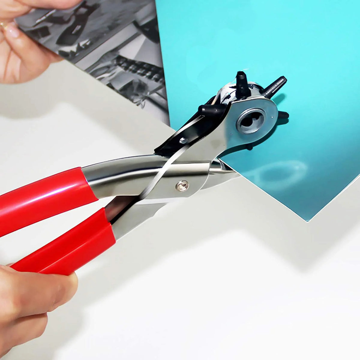A pair of red and silver leather hole punch pliers are shown gripping a piece of light blue paper.