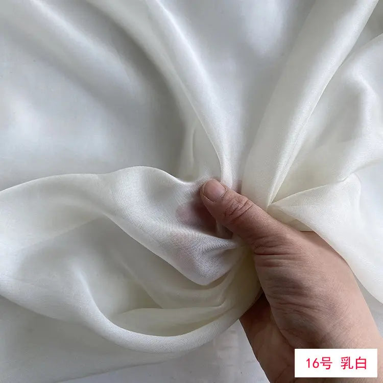 A hand is folding a piece of white silk chiffon fabric to show its drape and texture.