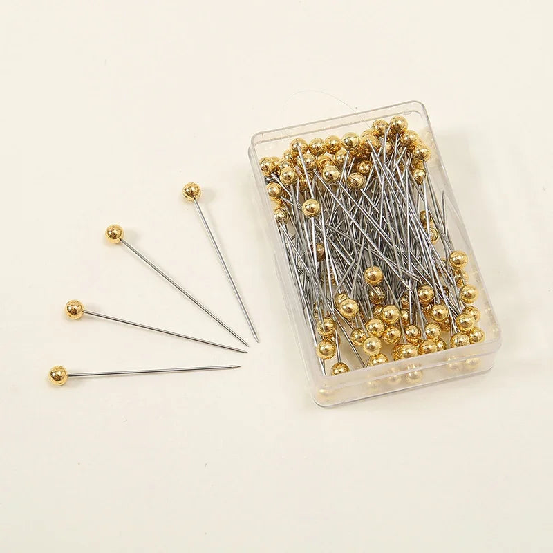 A clear plastic container filled with 100 gold-colored precision quilting pins for sewing and crafting.