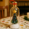 A clear glass bottle with a red cap holds a miniature Christmas tree and snow scene on a white base.
