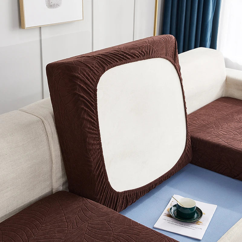 A close-up of a brown stretch jacquard sofa cover with a white cushion and blue accent fabric.