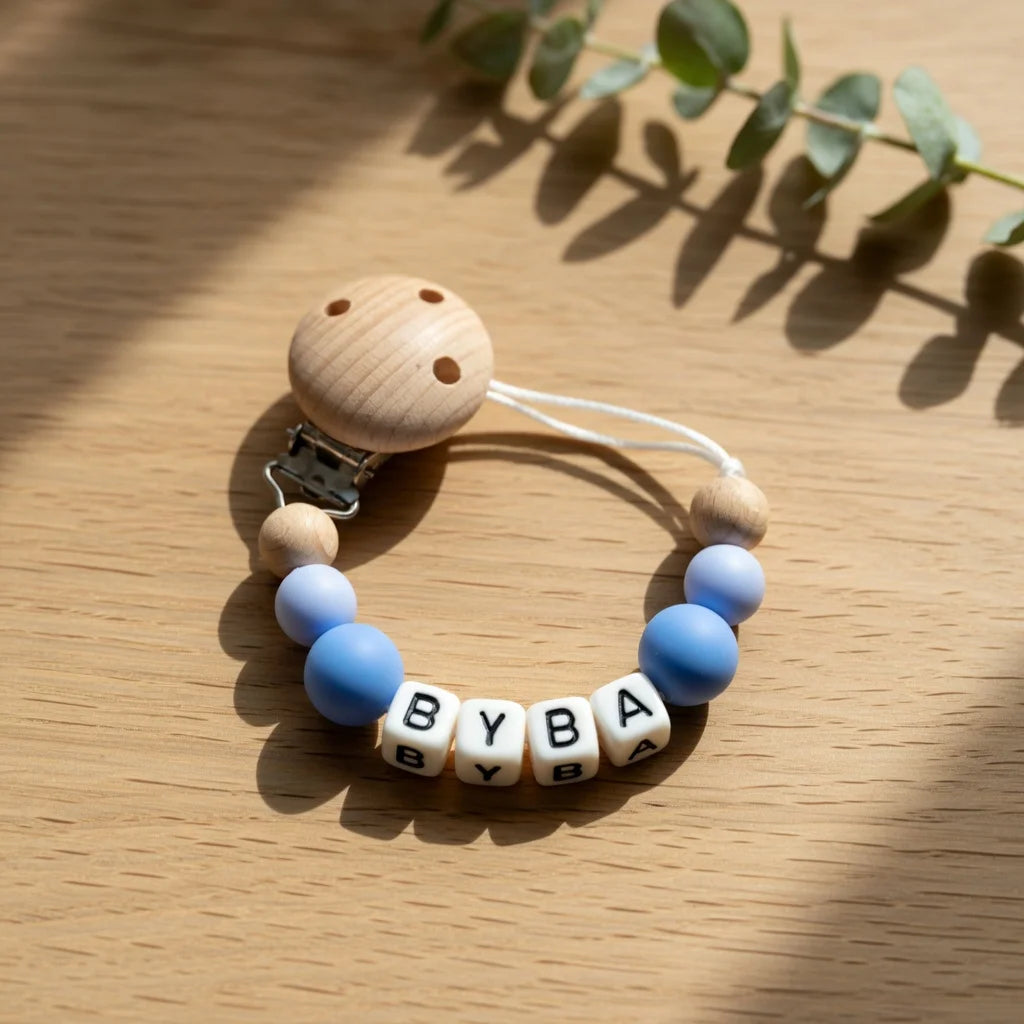 A wooden baby pacifier clip featuring custom lettering and a white cord, resting on a light wood surface.