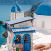 Hands hold a small blue wooden book nook kit with LED lighting in front of a white church building and sea view.