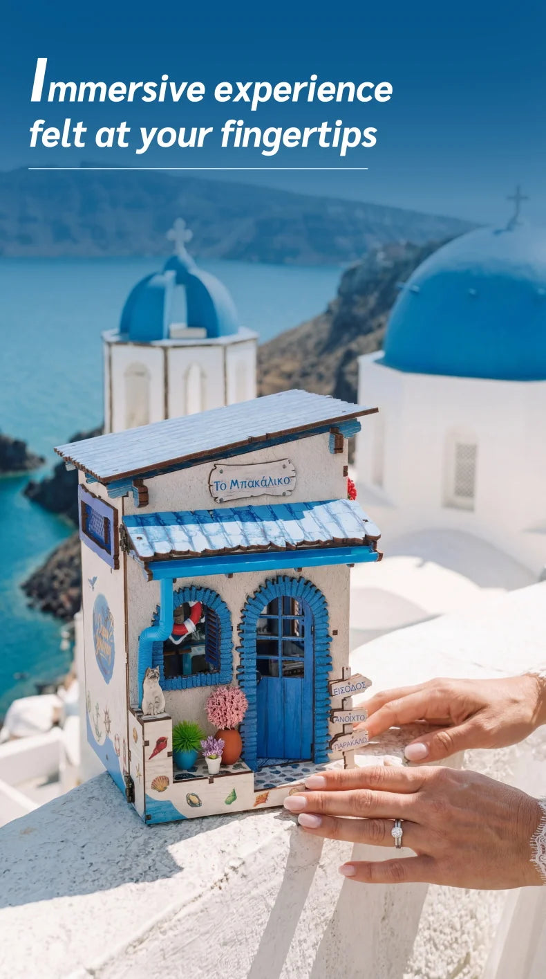 Hands hold a small blue wooden book nook kit with LED lighting in front of a white church building and sea view.
