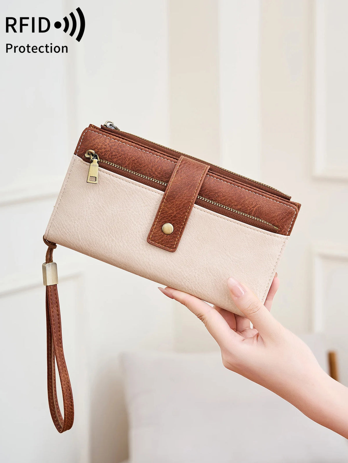 A hand holds a beige leather bifold wallet with a brown closure and strap, demonstrating RFID protection.