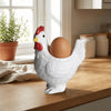 A white ceramic chicken figurine holding a brown egg, sitting on a wooden surface near a window.