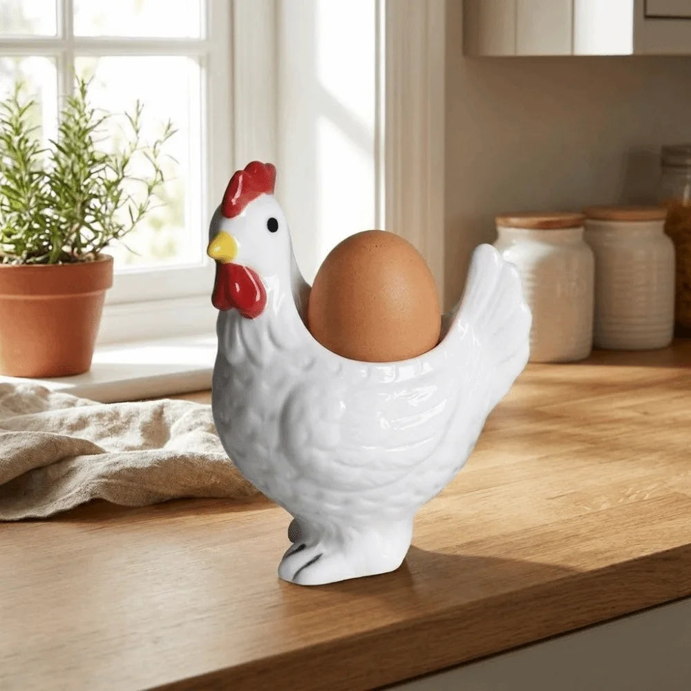 A white ceramic chicken figurine holding a brown egg, sitting on a wooden surface near a window.