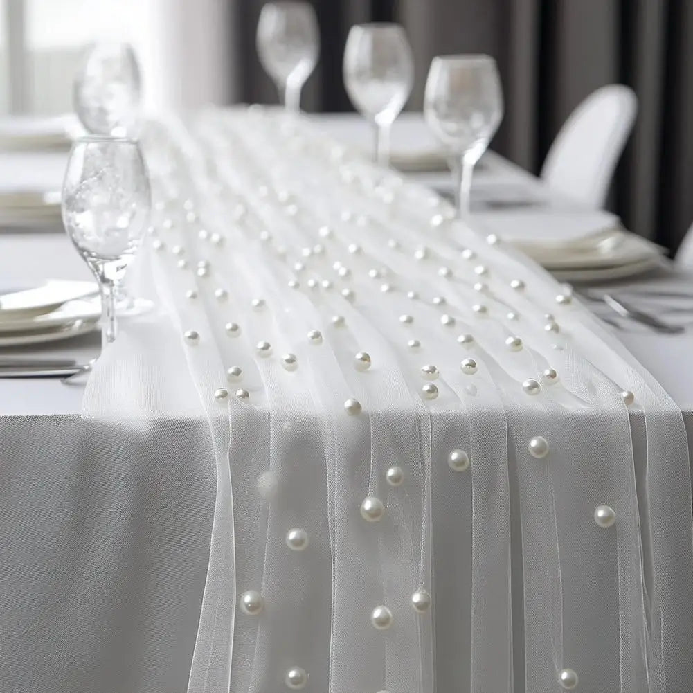 A white pearl beaded tulle table runner draped over a white tablecloth for an elegant wedding or event setting.