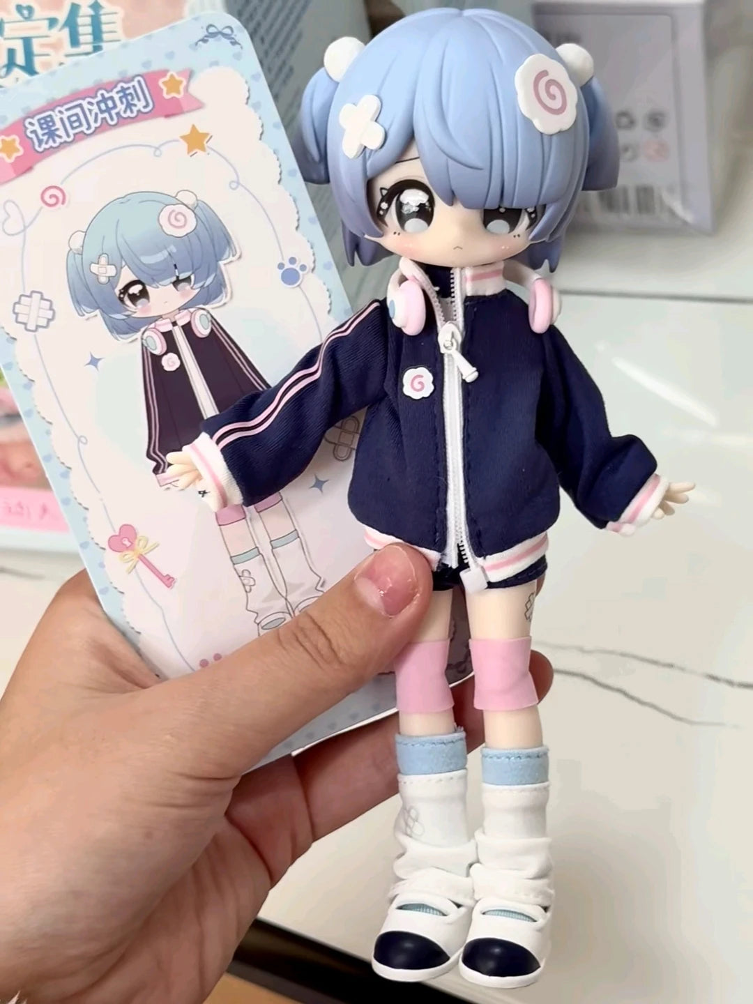 A hand holds an articulated BJD collectible figurine with light blue hair and a dark blue outfit.