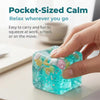 A hand is squeezing a translucent blue squishy ocean shell cube stress relief fidget toy on a white surface.
