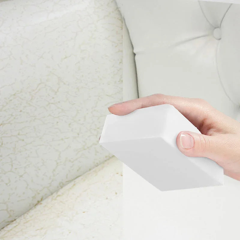 A hand holds a white melamine sponge cleaning eraser against a white upholstered surface.