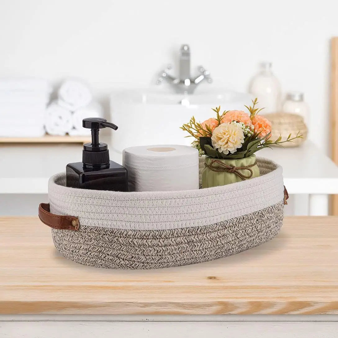 A woven cotton rope storage basket holding a roll of toilet paper and a small floral arrangement on a wooden surface.