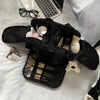 A black cosmetic travel organizer with multiple zippered compartments and a clear pouch is shown on a white surface.
