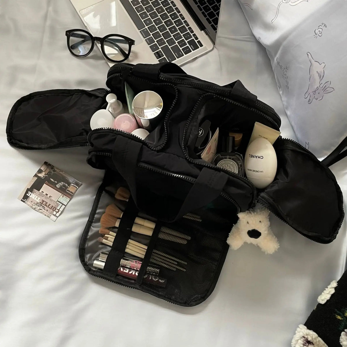 A black cosmetic travel organizer with multiple zippered compartments and a clear pouch is shown on a white surface.