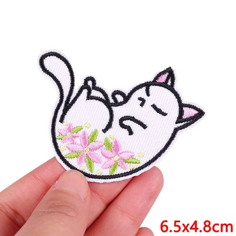 A hand holds a pink and white cartoon cat embroidery patch featuring closed eyes and floral details.