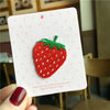 A white card featuring a red strawberry 3D embroidery patch, suggesting a gift or decorative item.