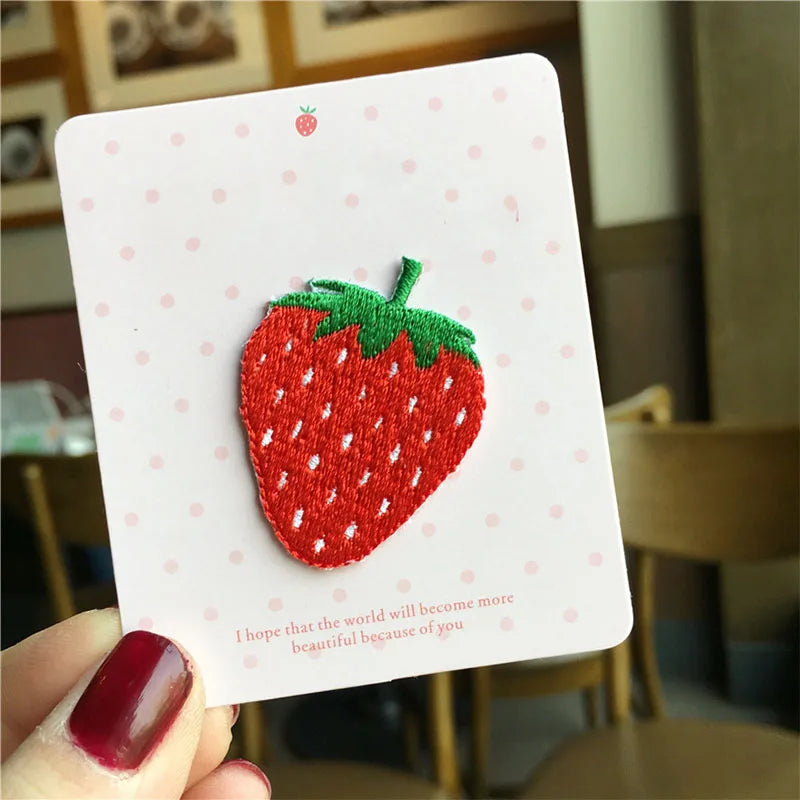 A white card featuring a red strawberry 3D embroidery patch, suggesting a gift or decorative item.