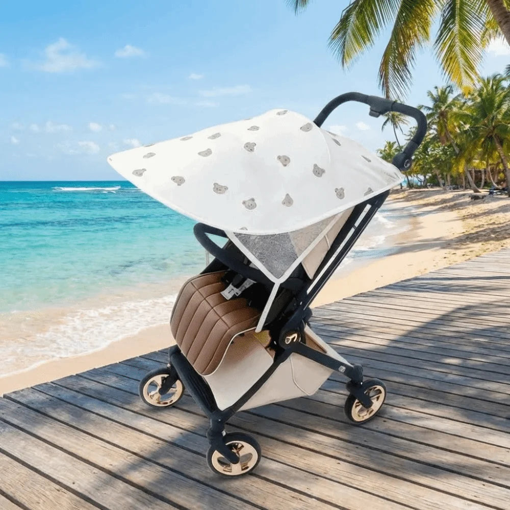 A beige and white stretchable UV-protective stroller sun shade is placed on a wooden deck near the ocean.