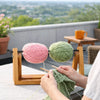 Hands are shown using a yarn ball holder to knit green yarn on a wooden table with a city view in the background.
