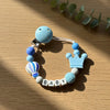 A light blue pacifier clip bracelet featuring round beads and a crown charm on a wooden surface.