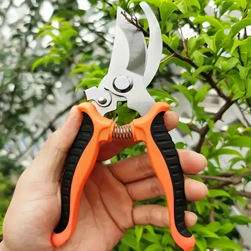 A hand holds a pair of orange and black stainless steel bypass pruning shears against a blurred green leafy background.