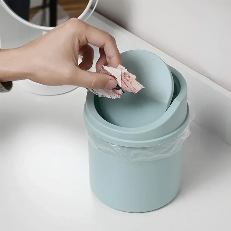 A hand is placing crumpled tissue paper into a light blue compact covered desktop trash bin.