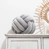 A gray knitted velvet throw pillow resting on a white wooden shelf next to a light-colored decorative object.