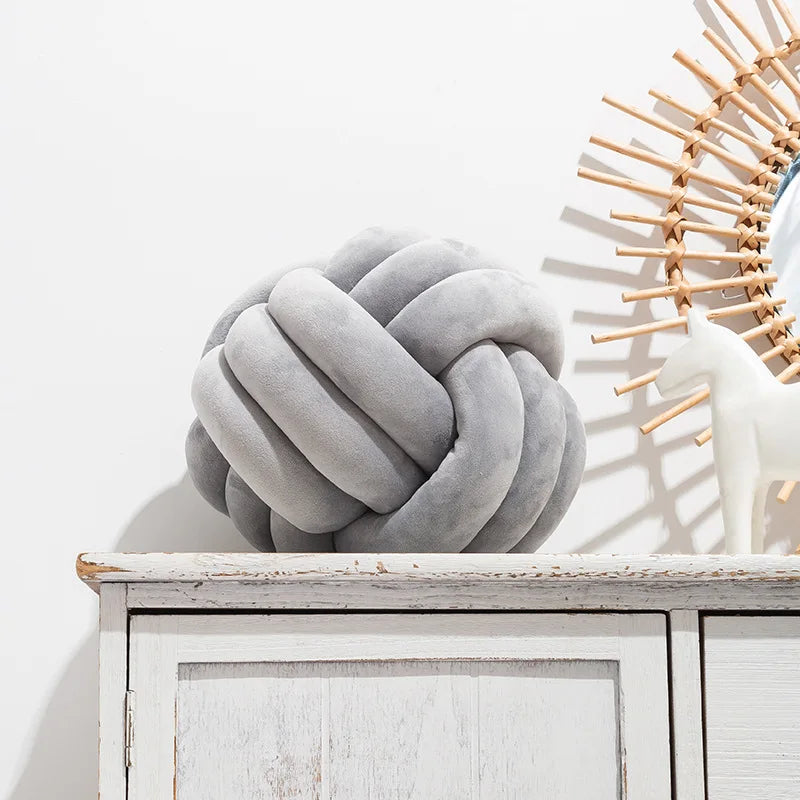 A gray knitted velvet throw pillow resting on a white wooden shelf next to a light-colored decorative object.