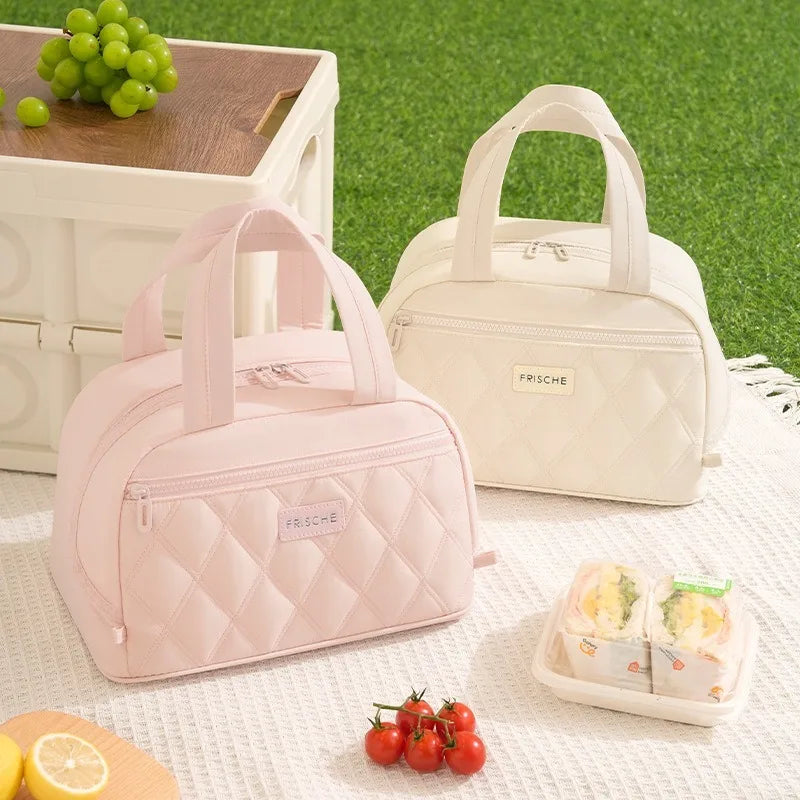 A pink quilted insulated lunch tote bag next to a cream-colored version and some fruit on a white surface.
