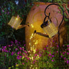 A vintage solar watering can garden light with a shepherd's hook illuminates plants in a garden setting.