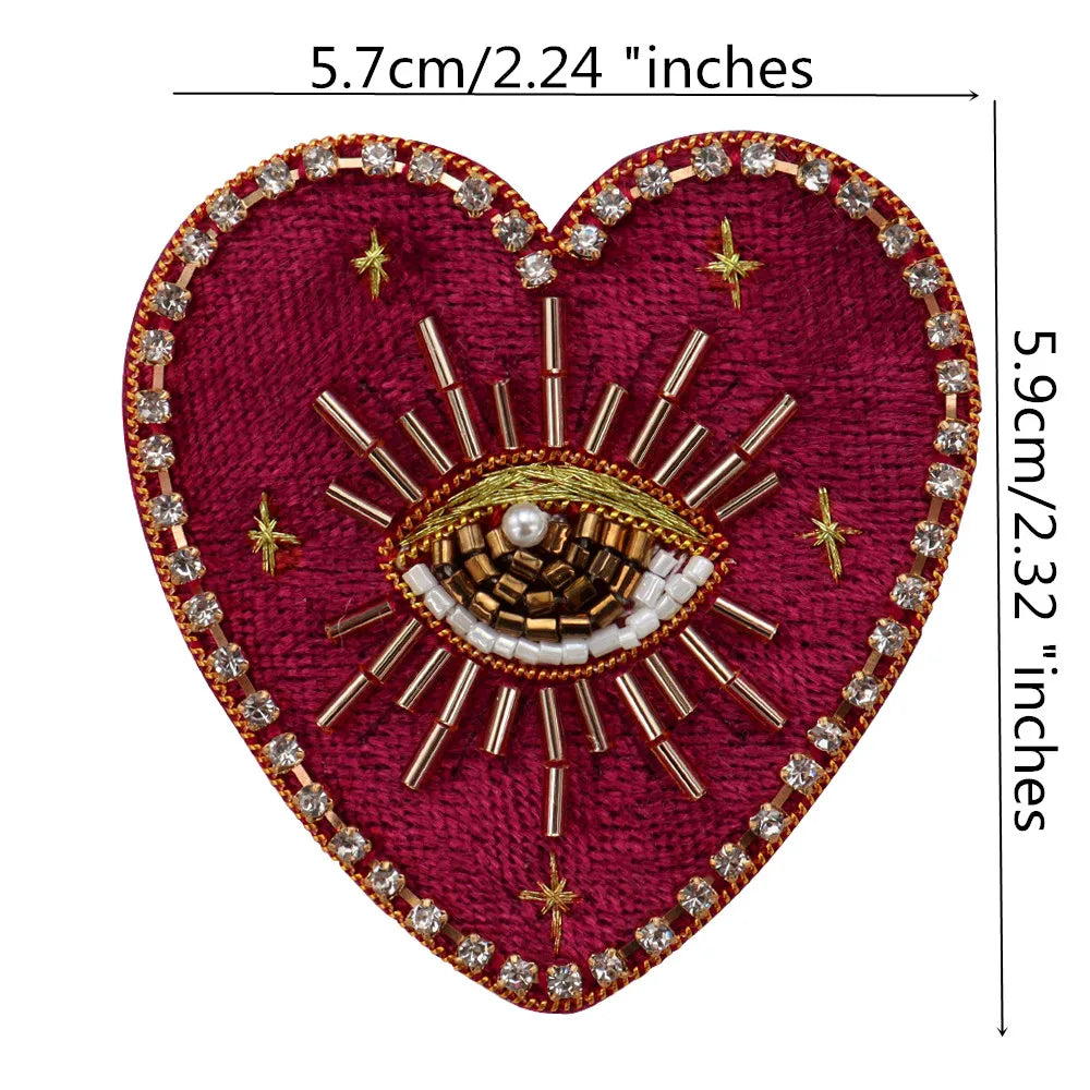 A deep magenta heart-shaped fabric patch decorated with gold and clear rhinestones forming an eye design.