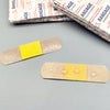 A strip of adhesive bandage with a yellow center is placed on a gray surface next to a stack of packaged bandages.