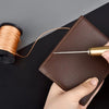 A hand holds a brown leather sewing awl set with a gold-colored needle and a spool of thread.