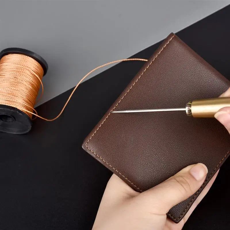 A hand holds a brown leather sewing awl set with a gold-colored needle and a spool of thread.