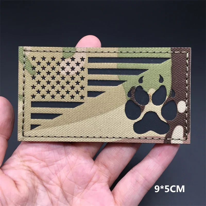 A rectangular tactical patch featuring a camouflage design with an American flag motif and paw print silhouette.