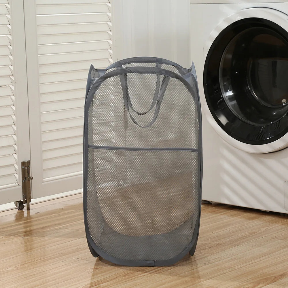 A gray foldable laundry hamper with a mesh design is shown next to a white washing machine.
